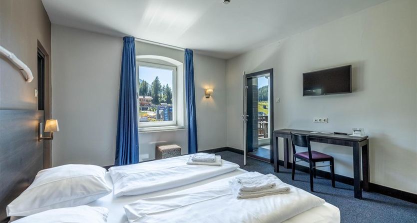 A modern hotel room with two beds and fresh white bed sheets. There is a table, a television, and a window overlooking the surroundings.
