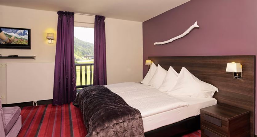A cozy hotel room with two beds and modern amenities. The walls are in purple and there is a balcony overlooking nature.