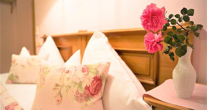 Een gezellige slaapkamer met witte kussens en een houten bed. Op het nachtkastje staat een vaas met verse rozen.