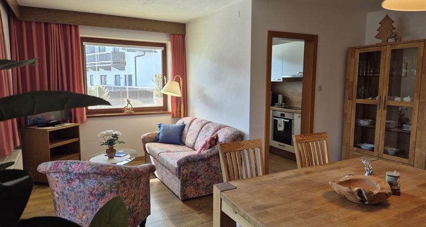 Ein gemütliches Wohnzimmer mit einem Sofa, zwei Sesseln und einem Esstisch. Der Raum hat große Fenster mit Blick nach draußen und ist hell und freundlich eingerichtet.