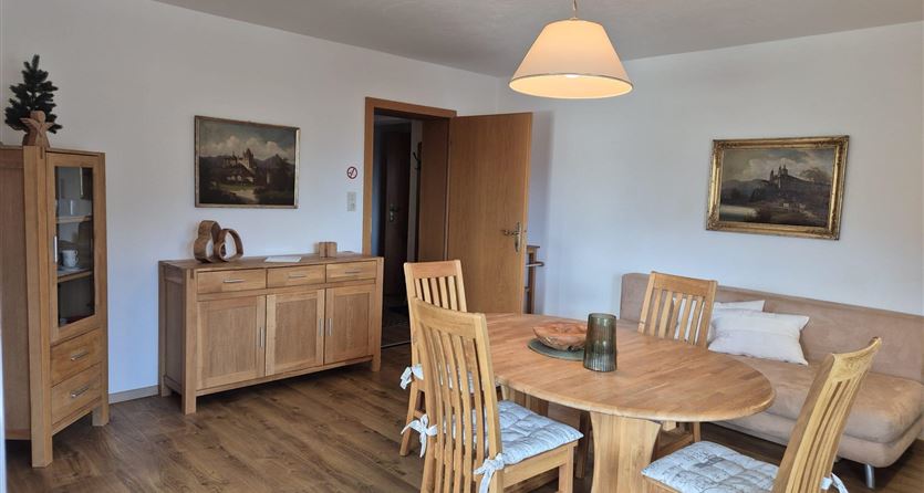 Ein gemütlicher Essbereich mit einem runden Holztisch und sechs Stühlen. An den Wänden hängen Bilder, und ein holzfarbener Schrank steht im Raum.
