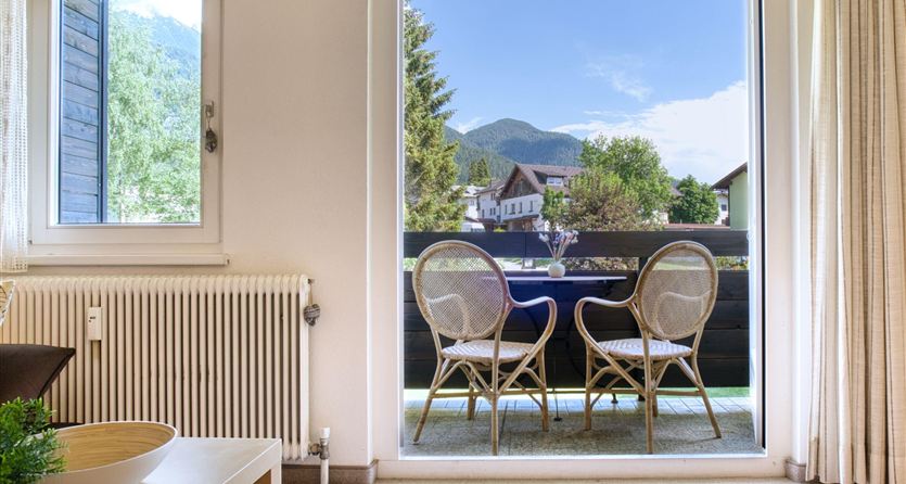 Una luminosa stanza con un balcone, da cui si può godere di una bella vista sul paesaggio. All'esterno ci sono due sedie e un tavolo, circondati da alberi e montagne.