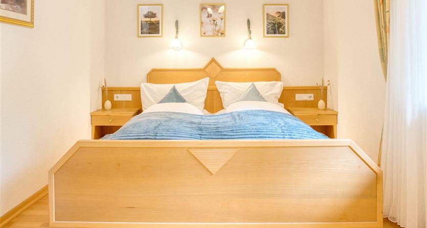 A cozy bedroom with a wooden bed and blue bedding. Pictures hang on the walls, and the light is soft and inviting.