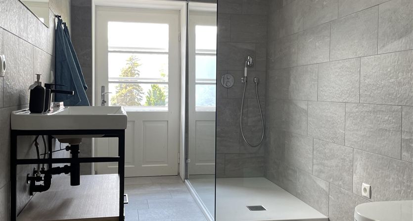 Ein modernes Badezimmer mit einer gläsernen Dusche und einem großen Fenster. Der Boden ist mit hellen Fliesen versehen und es gibt schwarze Badeteppiche.