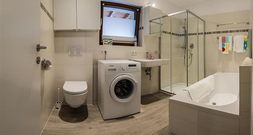 Ein modernes Badezimmer mit einer Dusche, einer Badewanne und einer Waschmaschine. Es hat helle Fliesen und ein Fenster für Tageslicht.