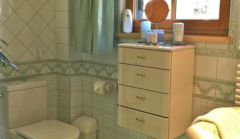 Ein Badezimmer mit weißen Fliesen und einem Fenster, durch das Licht scheint. Es gibt eine Toilette, ein Waschbecken mit einem Spiegelschrank und einen grünen Teppich.