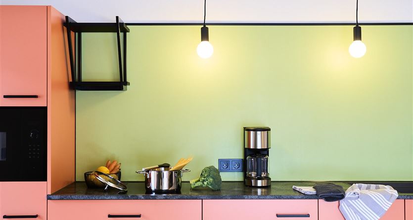 Eine moderne Küche mit korallenfarbener Schrankfront und grüner Wand. Auf der Arbeitsfläche stehen eine Kaffeekanne und frisches Obst.