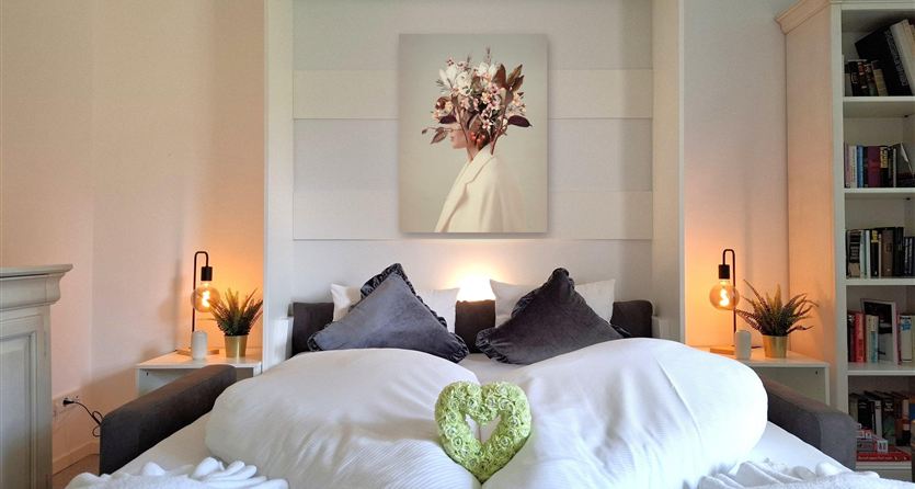 Een gezellig slaapkamer met een groot bed en decoratieve kussens. Aan de muur hangt een afbeelding van bloemen, en het licht creëert een ontspannen sfeer.