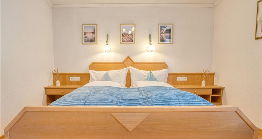 A cozy bedroom with a large double bed and white pillows. The walls are light and decorated with pictures, while night lamps and wooden dressers are added.