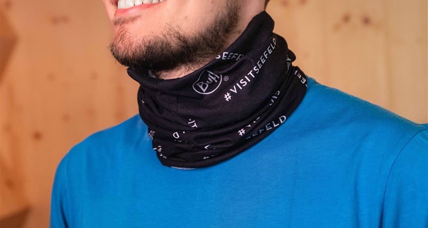 Ein Mann trägt einen schwarzen Schal mit Schriftzug. Er lächelt und trägt ein blaues T-Shirt, vor einem Holz-Hintergrund.
