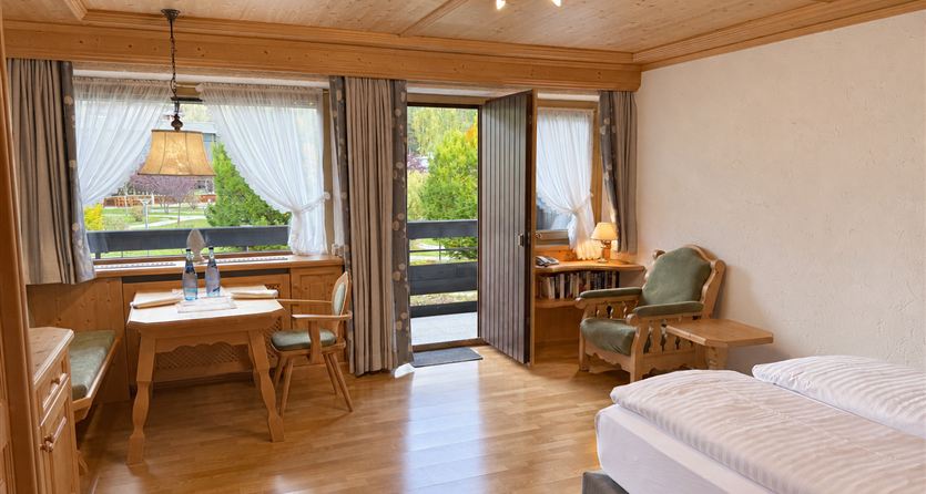 Ein gemütliches Zimmer mit Holzmöbeln und einem großen Fenster. Tisch und Stühle stehen neben einem Doppelbett, und der Blick öffnet sich zur Terrasse.