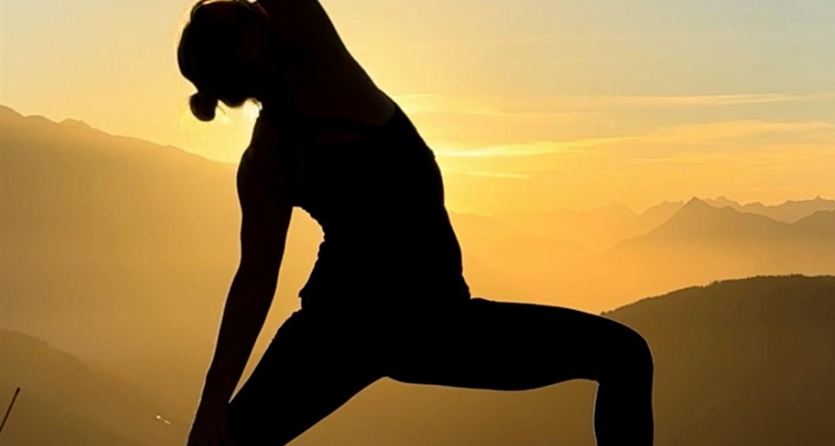 A person is doing yoga in a beautiful pose at sunset. In the background, there are mountains and a golden sky.