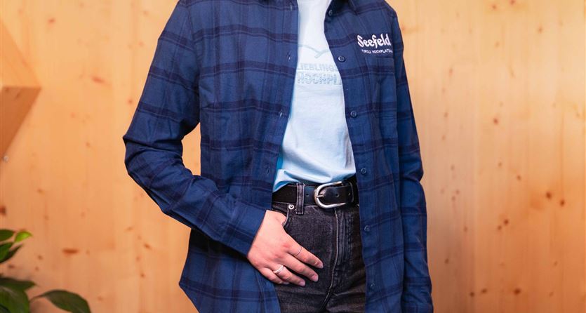 Ein Mensch trägt ein kariertes, blaues Hemd über einem hellblauen T-Shirt. Im Hintergrund ist eine Holzwand und ein Pflanze sichtbar.
