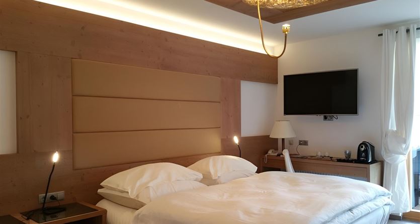 Een moderne slaapkamer met een tweepersoonsbed en houten accenten. Er is een televisie aan de muur en grote ramen die voor daglicht zorgen.