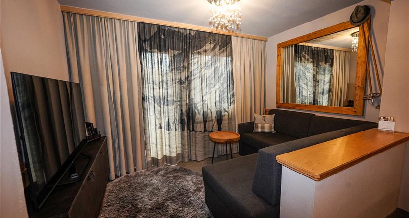 Ein gemütliches Wohnzimmer mit einem grauen Sofa, einem kleinen Tisch und einem Fernseher. Große Vorhänge lassen viel Licht herein und ein Spiegel vergrößert den Raum.