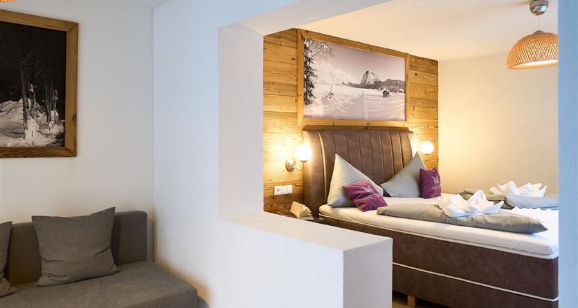 A cozy hotel room with a large bed and wooden details. A seating area with a gray sofa complements the space.