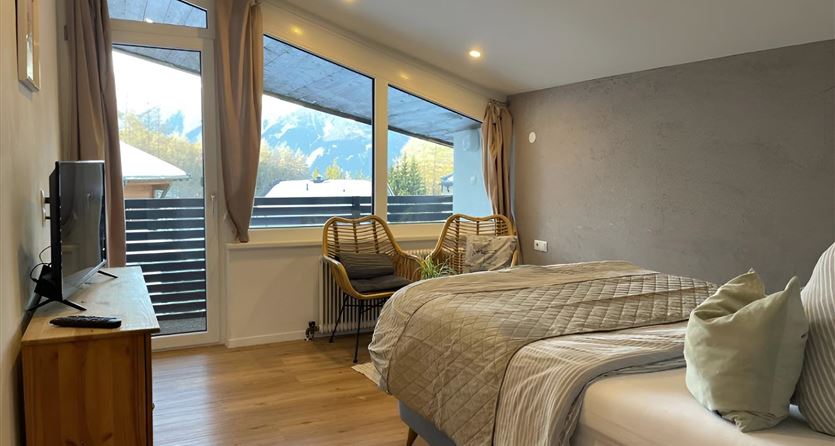 Ein modernes Schlafzimmer mit einem großen Bett und zwei gemütlichen Stühlen. Große Fenster bieten einen Blick auf die Berge und bringen viel Tageslicht hinein.