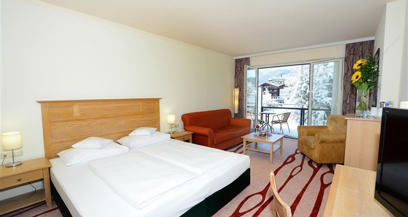 Een moderne hotelkamer met een groot bed, een gezellige fauteuil en een balkon. Door de ramen is een met sneeuw bedekt landschap zichtbaar.