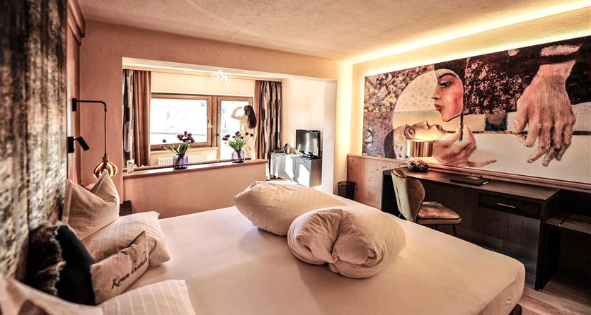 Ein modernes Hotelzimmer mit einem großen Bett und künstlerischer Wandgestaltung. Es gibt Fenster mit Blick nach außen und stilvolle Möbel.