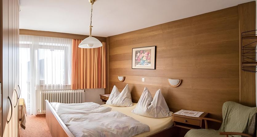 Ein gemütliches Schlafzimmer mit einem Doppelbett und zwei Kopfkissen. Der Raum hat Holzwände, einen Sessel und große Fenster mit Vorhängen.