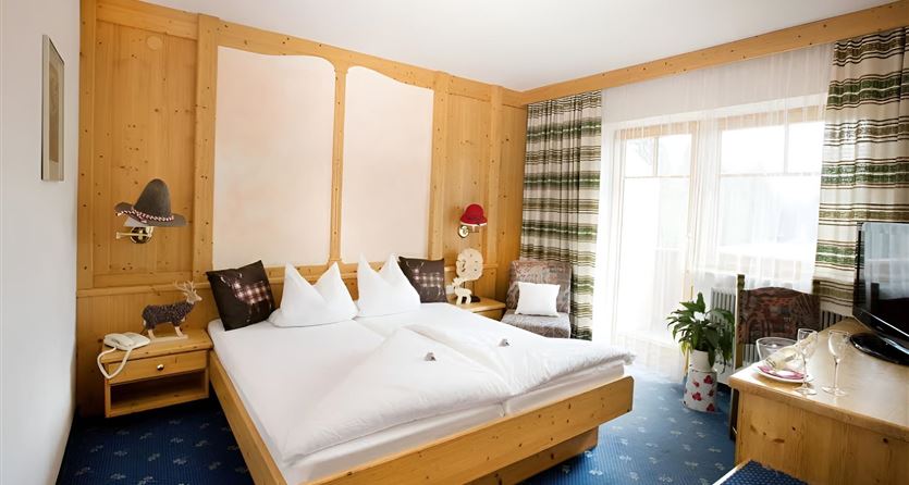 Ein gemütliches Hotelzimmer mit einem Doppelbett und einem Holzdesign. Große Fenster lassen viel Tageslicht herein und es gibt einen kleinen Tisch mit Stühlen.