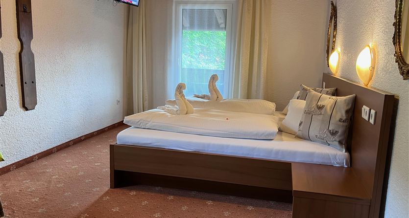 Een gezellig slaapkamer met een tweepersoonsbed en zachte kleuren. Een televisie hangt aan de muur, en er zijn grote ramen die daglicht binnenlaten.