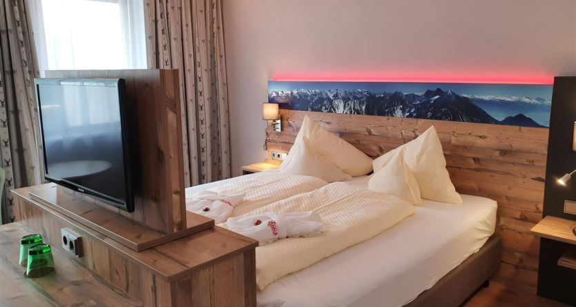 Ein modernes Schlafzimmer mit einem großen Bett und weichen Kissen. Ein Fernseher steht auf einem Holzmöbel und es gibt eine gemütliche Atmosphäre durch die indirekte Beleuchtung.