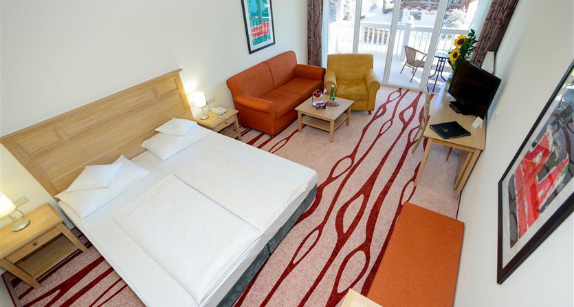 Ein gemütliches Hotelzimmer mit einem Doppelbett, einem Sofa und einem Fernseher. Große Fenster bieten Zugang zu einem Balkon mit Aussicht.