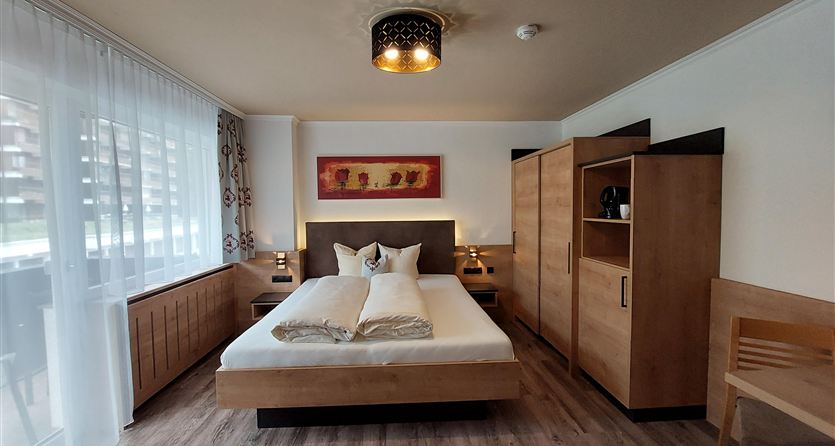 Een gezellige slaapkamer met een groot bed en houten meubels. Er zijn ramen met gordijnen die veel licht binnenlaten.