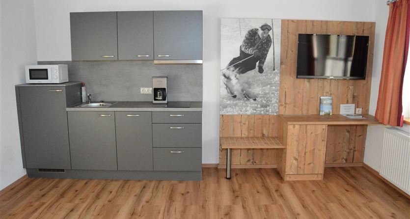 Een moderne keuken met grijze kasten en houten planken. In de kamer staat een televisie en een groot schilderij aan de muur.