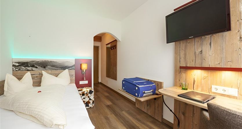 Ein modernes Hotelzimmer mit einem Doppelbett und einem Schreibtisch. An der Wand hängt ein Fernseher und eine blaue Tasche steht neben dem Bett.
