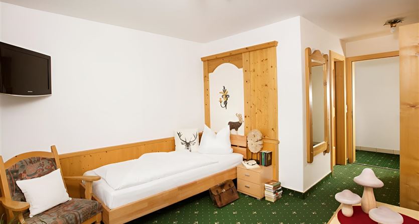 Ein gemütliches Zimmer mit einem Holzbett, einem Sessel und einem Fernseher an der Wand. Der Boden ist mit grünem Teppich ausgelegt, und es gibt rustikale Dekorationselemente.