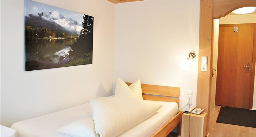 A simple room with a single bed and large cushions. On the wall hangs a picture of a picturesque landscape with a lake and mountains.