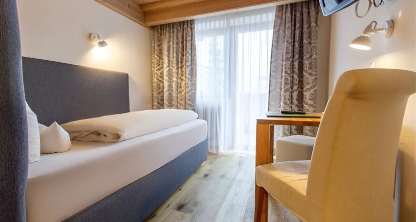 A modern hotel room with a single bed and a desk. The large window front lets in a lot of light and creates a bright atmosphere.