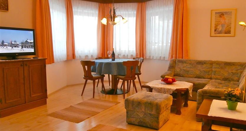 A cozy living room with a corner couch and a table. There are large windows with curtains and a television in the room.