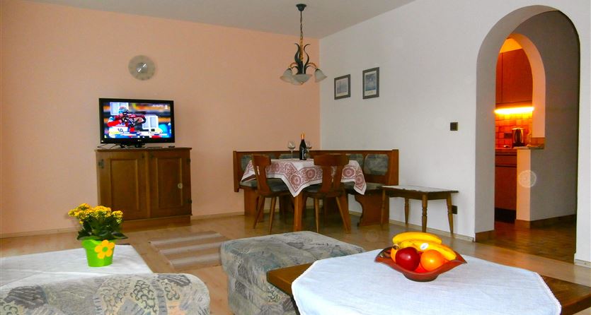 A cozy living room with a television, a dining area, and an open kitchen. A fruit bowl and flowers on the table create an inviting atmosphere.