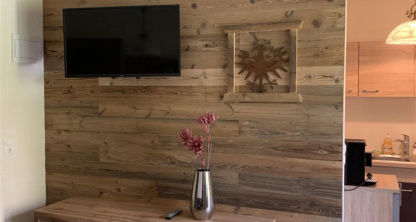 Een elegante woonkamer met een houten bekleding en een televisie aan de muur. Een houten tafel met decoratieve elementen en een open keuken op de achtergrond.