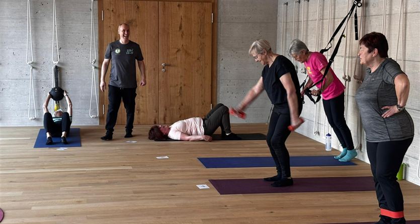Eine Gruppe von Menschen trainiert in einem Fitnessraum. Einige üben auf Matten, während andere mit Fitnessgeräten arbeiten.
