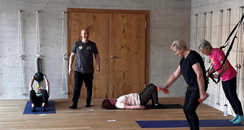 A group of people is doing gymnastics in a bright room. Some are using mats, while others are training with weights.