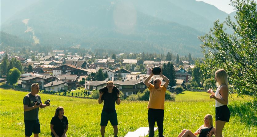 Eine Gruppe von Menschen trainiert auf einer grünen Wiese mit Bergen im Hintergrund. Die Stadt ist im Tal sichtbar, und das Wetter ist sonnig.