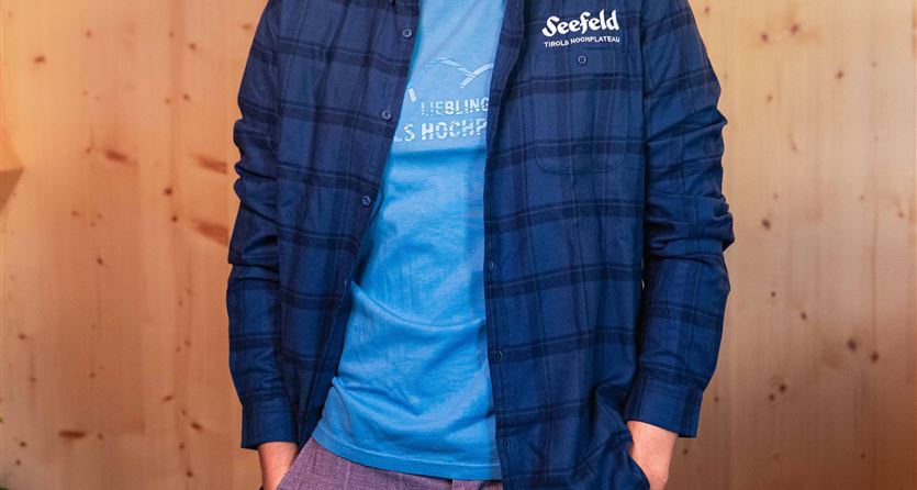 Ein junger Mann trägt ein kariertes Hemd und ein T-Shirt in blauer Farbe. Der Hintergrund ist aus Holz und vermittelt eine gemütliche Atmosphäre.