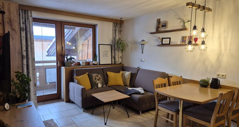 Een gezellige woonkamer met een bank, een tafel en een eetgedeelte. Er zijn ramen met uitzicht en warm licht door de hanglampen.