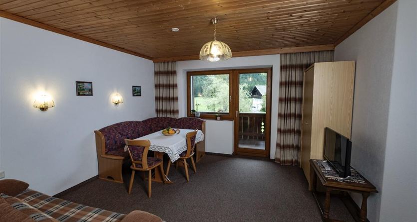 Ein gemütlicher Wohnraum mit Holzwänden und einem großen Fenster. Es gibt eine Sitzecke mit einem Tisch und Stühlen sowie einen Fernseher.