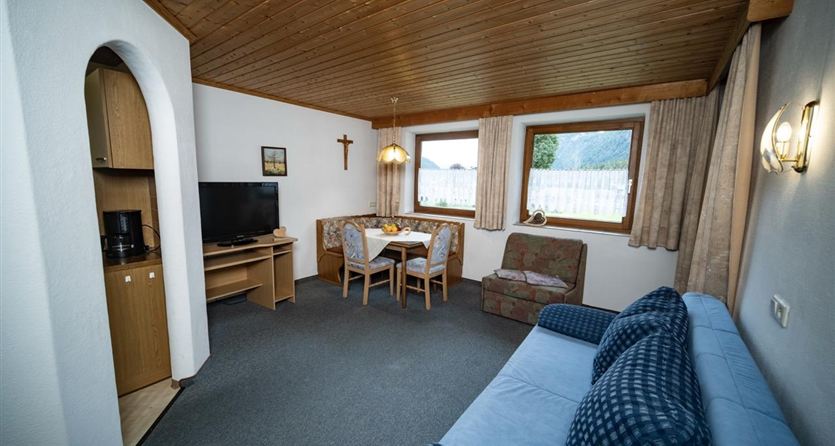 Ein gemütlicher Wohnraum mit einer blauen Couch, einem Esstisch und einem Fernseher. Die Fenster bieten einen Blick nach draußen, und die Holzbalkendecke sorgt für eine warme Atmosphäre.