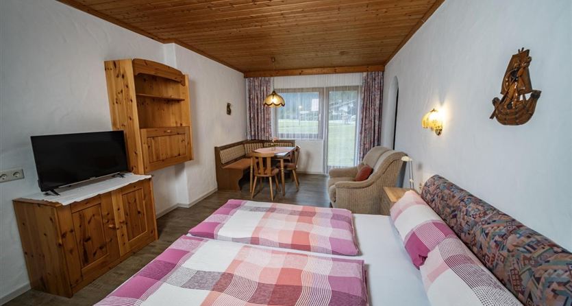 Ein gemütliches Zimmer mit zwei Betten, einem Fernseher und einem Essbereich. Große Fenster sorgen für viel Tageslicht und einen Blick nach draußen.