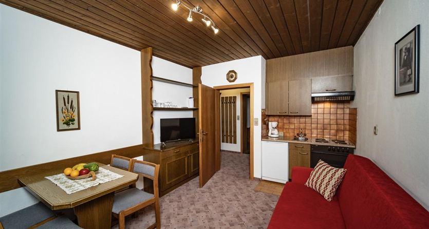 Ein gemütliches Wohnzimmer mit einer roten Couch, einem Esstisch und einer kleinen Küche. Die Wände sind hell und das Holzdecke verleiht dem Raum eine warme Atmosphäre.