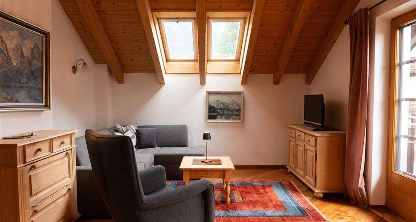 Ein gemütliches Wohnzimmer mit Holzdecken und Fenstern. Es gibt ein Sofa, einen Tisch und einen Fernseher in einem einladenden Raum.