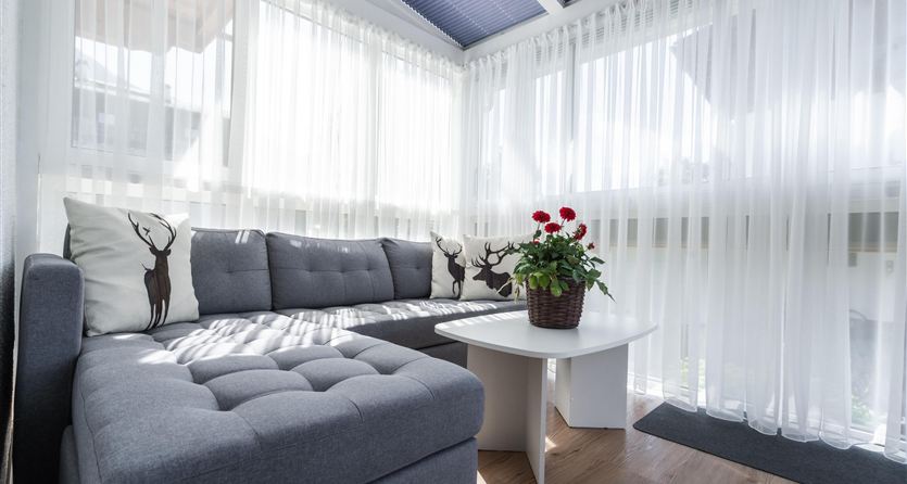 Ein gemütlicher Wohnbereich mit einem grauen Sofa und einem kleinen weißen Tisch. Große Fenster mit Vorhängen lassen viel Licht herein.