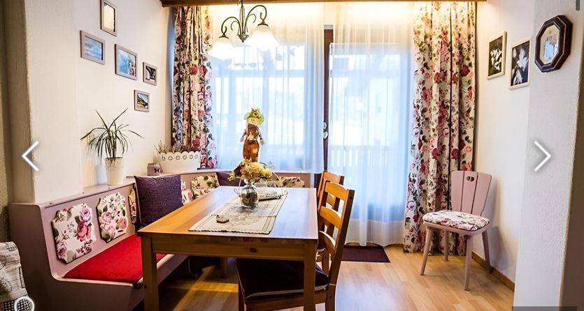 Ein gemütlicher Essbereich mit einem Holztisch und bunten Stühlen. Die Wände sind mit Blumenmustern und Bildern dekoriert, und das Licht strömt durch die Fenster.