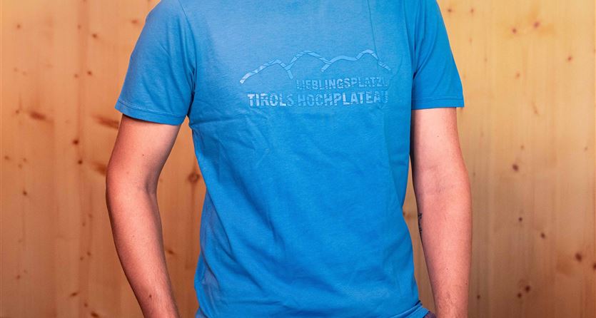 Ein junger Mann trägt ein blaues T-Shirt mit dem Schriftzug „Lieblingsplatz Tirols Hochplateau“. Im Hintergrund ist eine Holzverkleidung zu sehen.